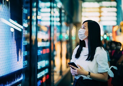 Economic and financial impact during the Covid-19 health crisis deepens. Businesswoman with protective face mask checking financial trading data on smartphone by the stock exchange market display screen board in downtown financial district showing stock market crash sell-off in red colour