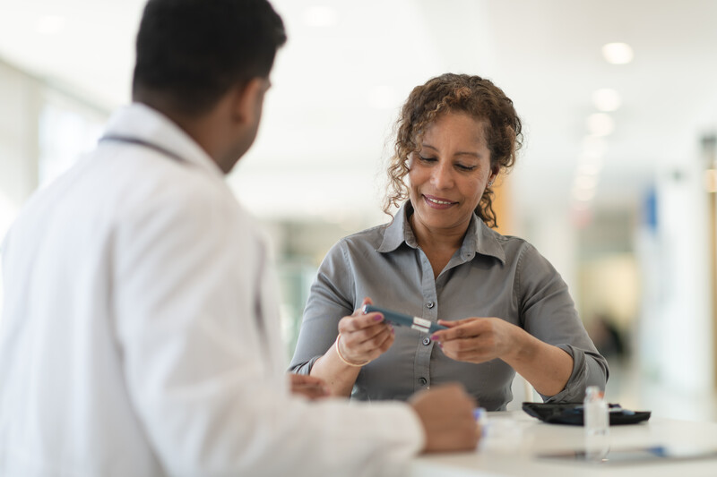 iStock-1146576913 A male Indian doctor meets with a female patient. The woman is a mature adult of African descent. The two people are seated at a table. The patient is diabetic and is holding an insulin pen. The doctor is explaining how to use the device.
