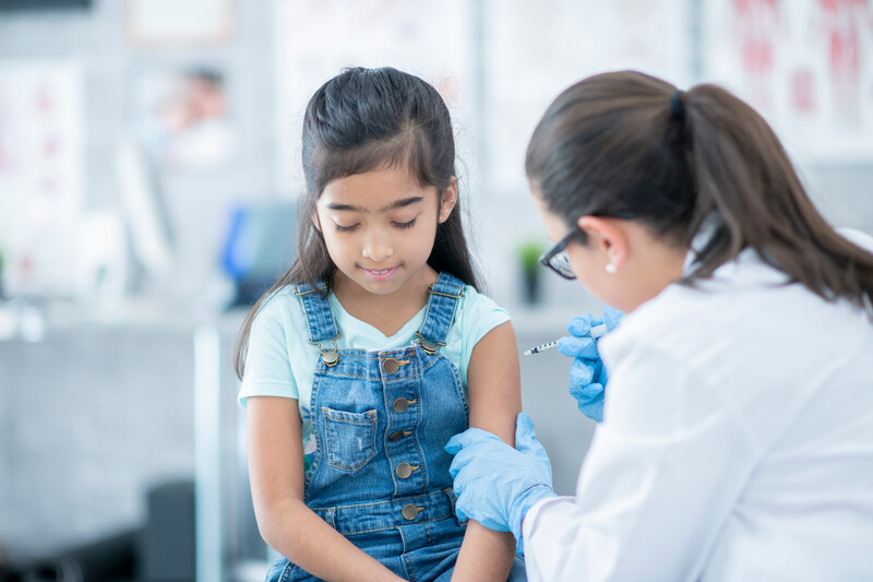 iStock-1065521378 A little girl gets a vaccination from her doctor. She looks down as she wears a brave face. Her doctor is holding her arm.
